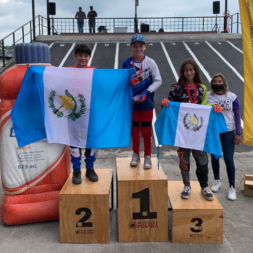 Descripción de la foto para personas con discapacidad visual: Niños sostienen con ambas manos sus banderas en el podio de premiación del evento. (Créditos: FGC)