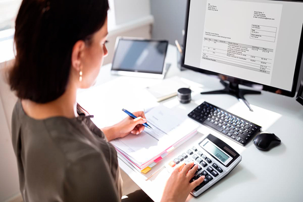 Cómo tramitar documentos tributarios en línea, Guatemala - Descripción de foto - mujer haciendo cuentas en una calculadora, mientras anota y observa la computadora con una factura