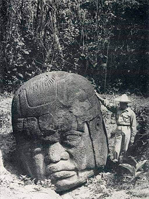 Descripción de foto - Arqueólogo a la par de una cabeza colosal Olmeca tallada en piedra, a blanco y negro. - Crédito de foto - Laura S