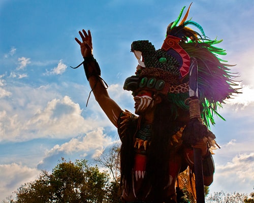 Descripción de foto - Hombre vestio de guerrero maya en el atardecer, levantando su mano. - Crédito de foto - Alberto Ramírez Gordillo