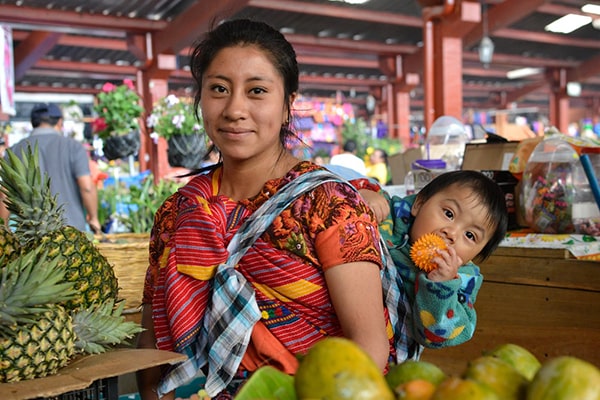 Descripción de foto - Mujer indígena guatemalteca, en el mercado, llevando en su espalda a su hijo. - Crédito de foto - Ministerio de Trabajo