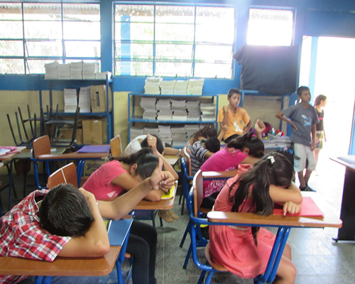 Descripción de la foto para personas con discapacidad visual - Niños en un salón recostados en sus pupitres boca abajo con el dedo levantado. - Crédito de foto - Chicos and Kids Inc
